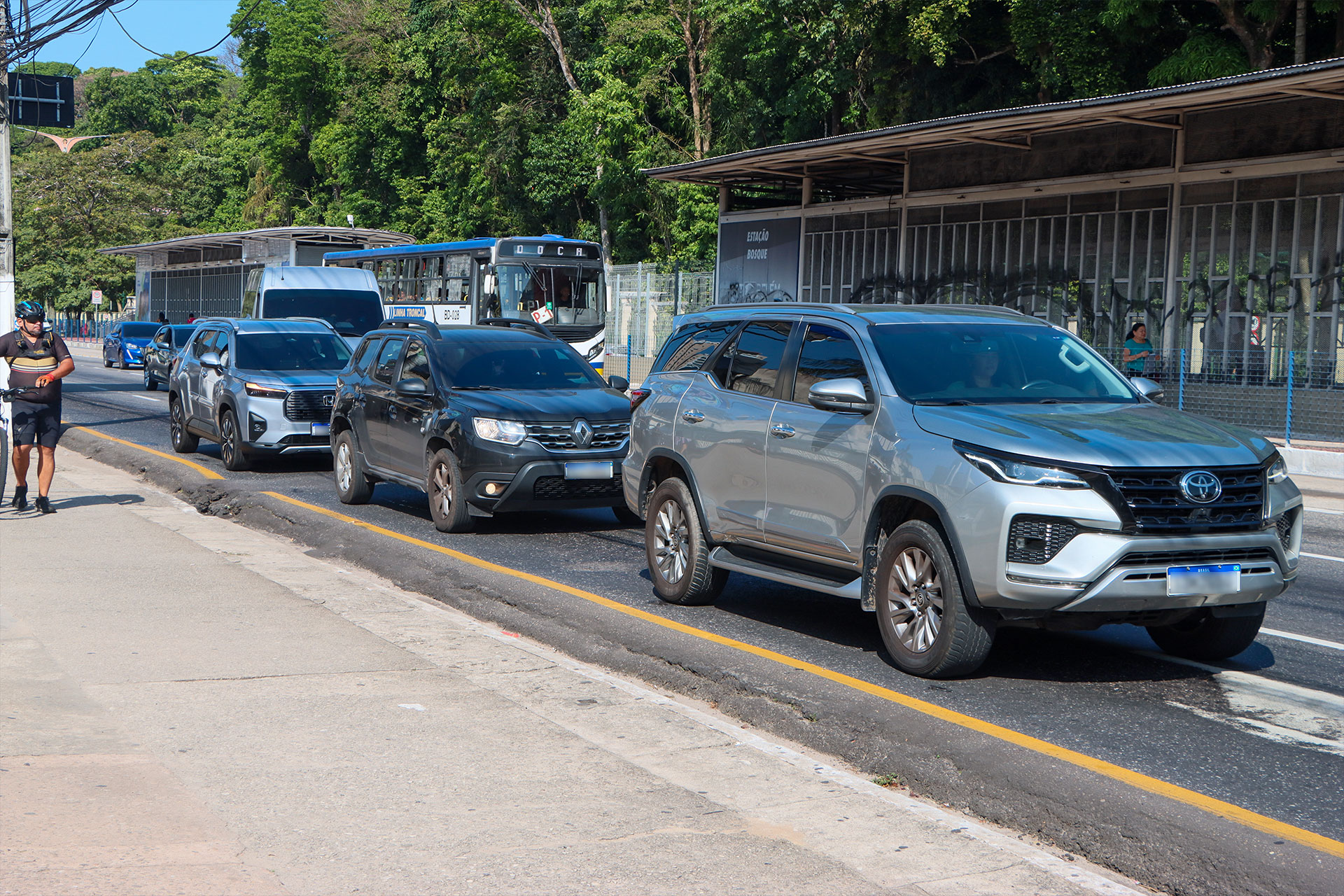 Fila de SUVs ocupa a faixa junto à Estação Bosque do BRT em Belém, enquanto um ciclista pedala espremido no canteiro e um ônibus espera atrás
