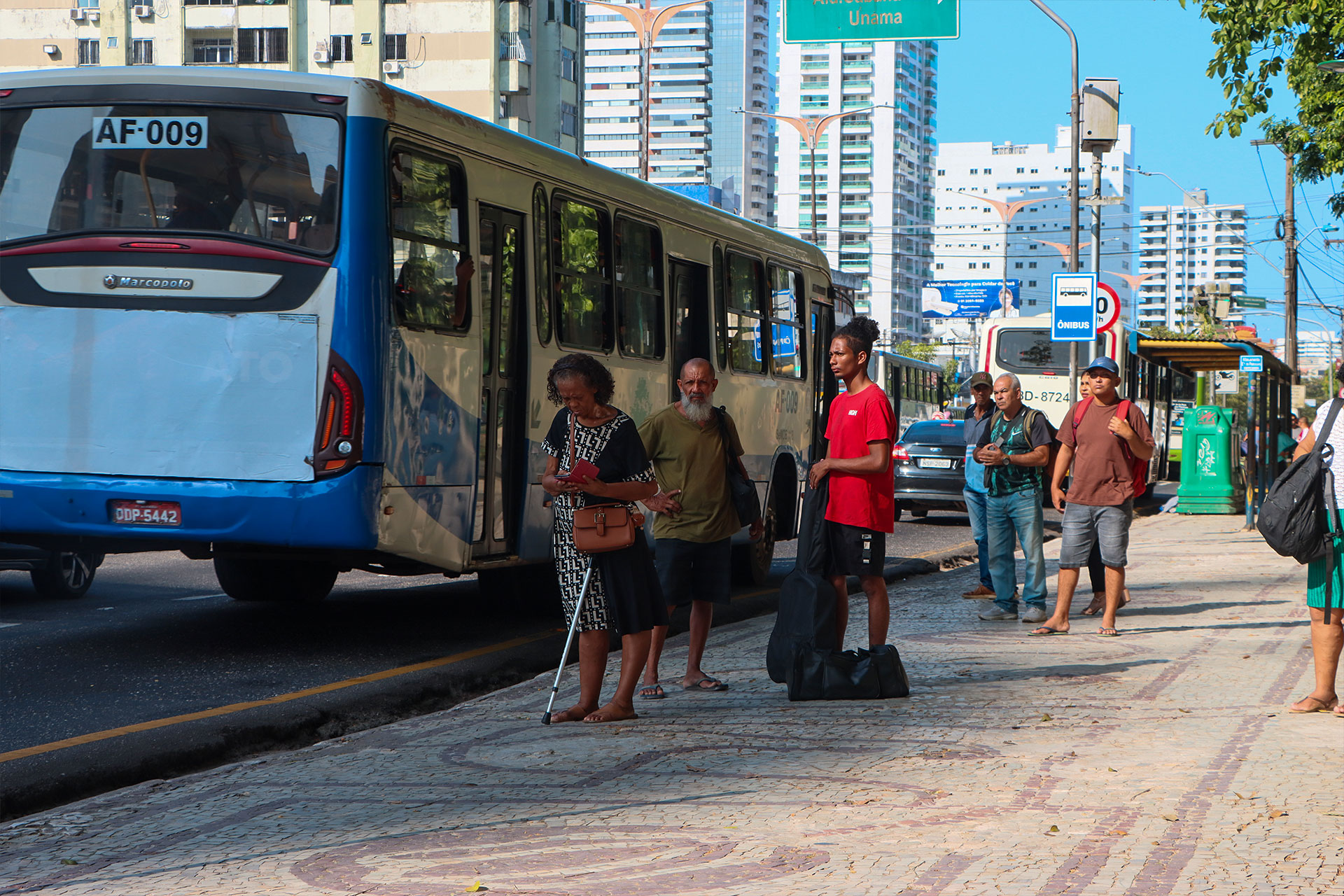 Passageiros esperam em pé na calçada de pedra portuguesa ao lado de um ônibus urbano parado em avenida de Belém